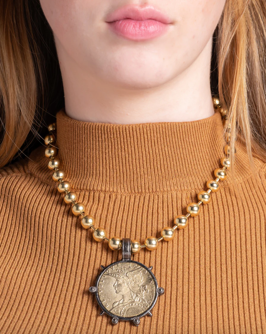 A person wearing a turtleneck sweater with a coin necklace that has a gunmetal outline and appears to be gold-plated over brass.