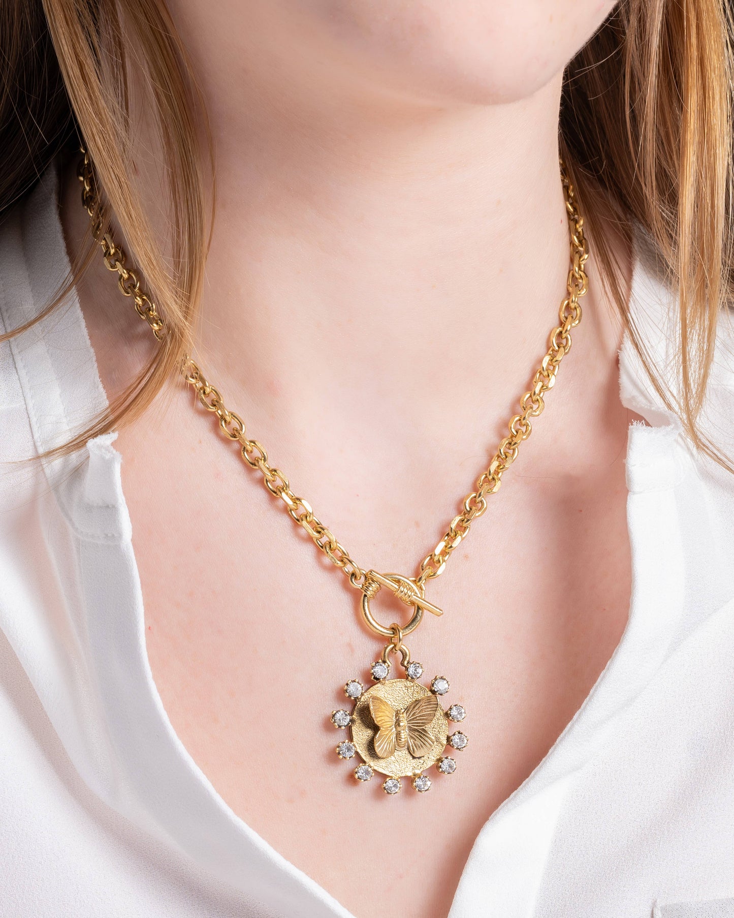 A gold-plated necklace with a circular pendant featuring a butterfly design, worn by a person with a white shirt.
