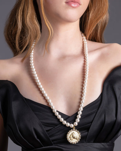 A person wearing a black off-shoulder top and a 22K gold plated over brass necklace with a pearl strand and a gold medallion.